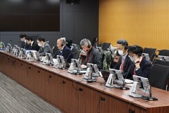 写真:会議の様子1(机上に一人1台タブレットが設置されている長机に座っている参加者たち。)諏訪sるいながつくえに)