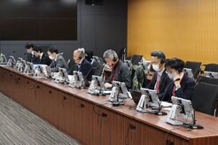 写真：会議の様子1（机上に一人1台タブレットが設置されている長机に座っている参加者たち。）諏訪sるいながつくえに）